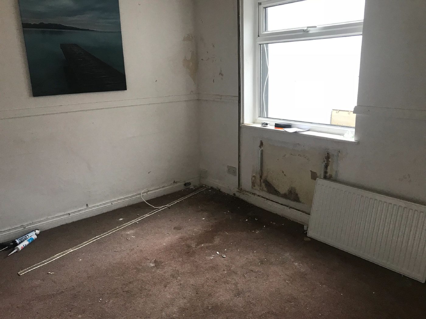 A room with blown, stained plaster below the window and behind the radiator, the damage stopping about a metre up the wall