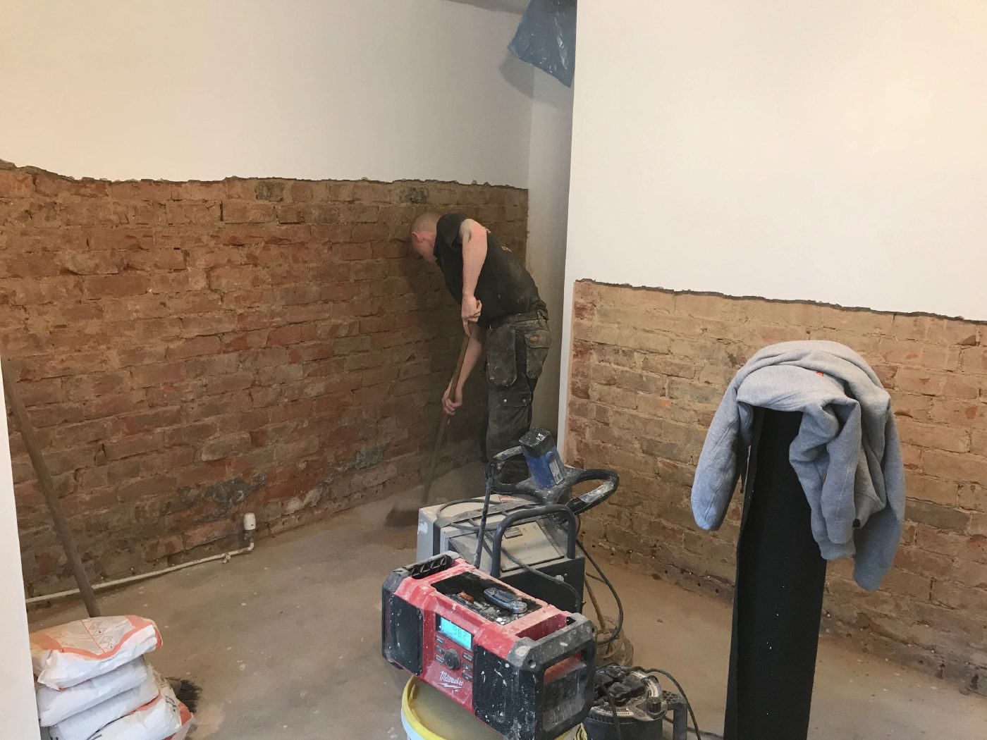 Salt-contaminated plaster stripped back to bare brickwork around two walls of a room, with a member of the team clearing the base of the wall