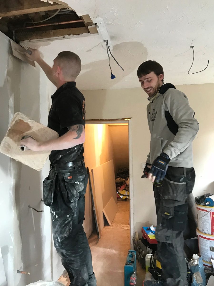 Two of the team working in a house, one skimming a wall by a doorway while the other looks on